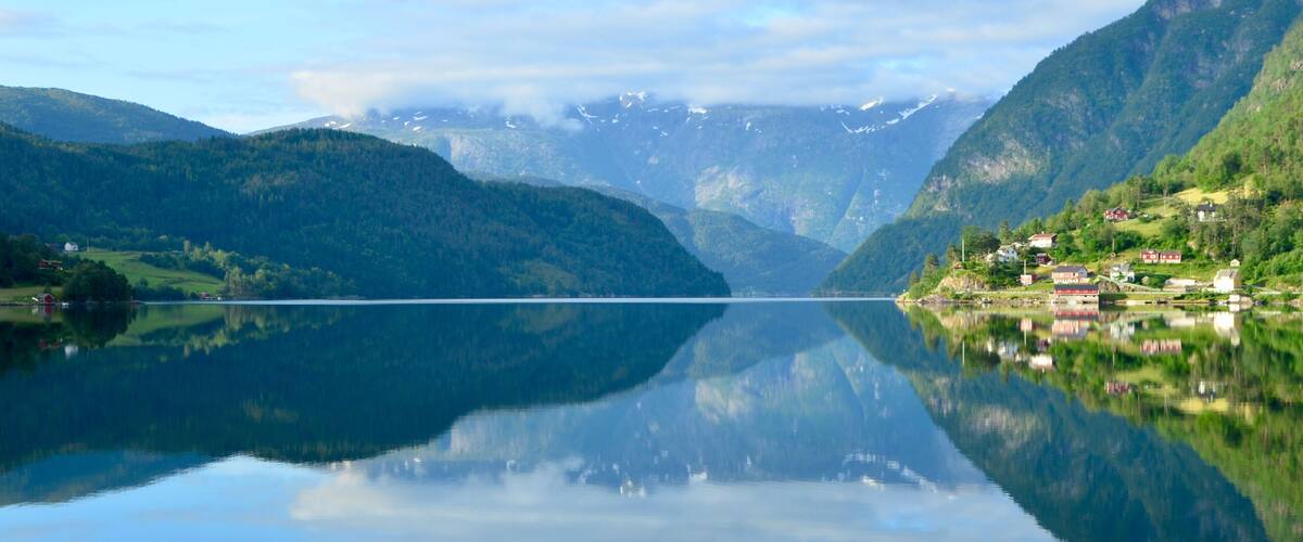 A calm lake with a clear reflection, surrounded by green hills and a few houses.