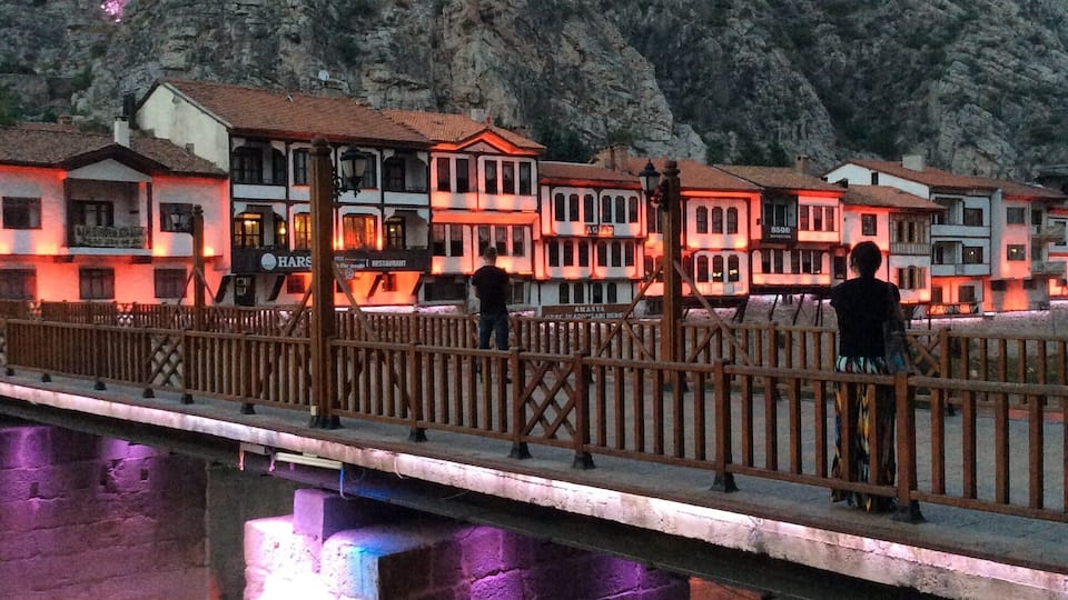 A stone bridge over a river with buildings on the opposite bank, illuminated by purple lights.