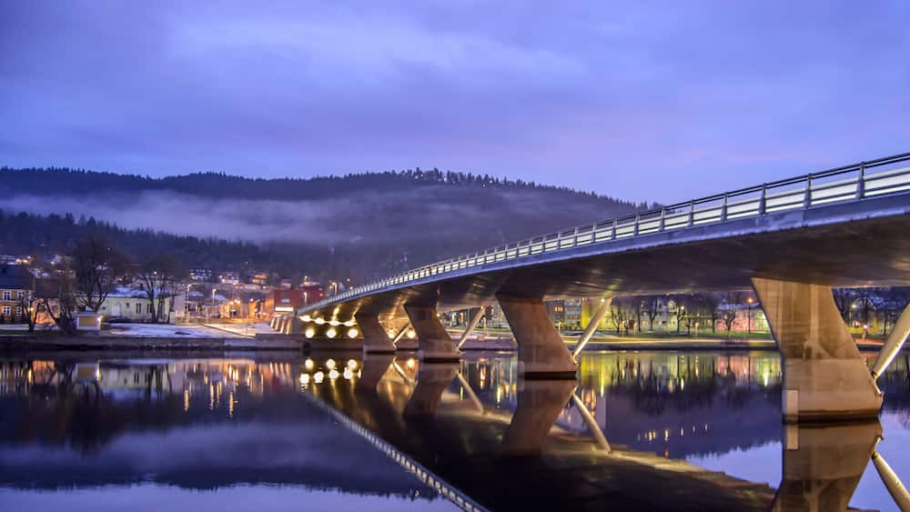Un pont enjambant une rivière paisible, reflétant les lumières d’une ville, avec une colline enveloppée de brume en arrière-plan.