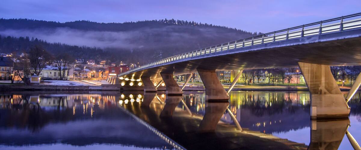 A bridge over a calm river reflecting the lights of a town and a mist-covered hill in the background.