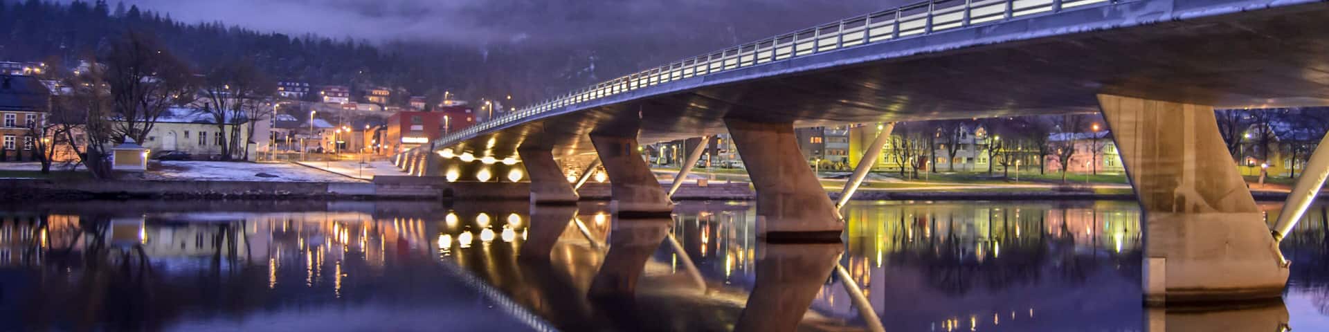 A bridge over a calm river reflecting the lights of a town and a mist-covered hill in the background.