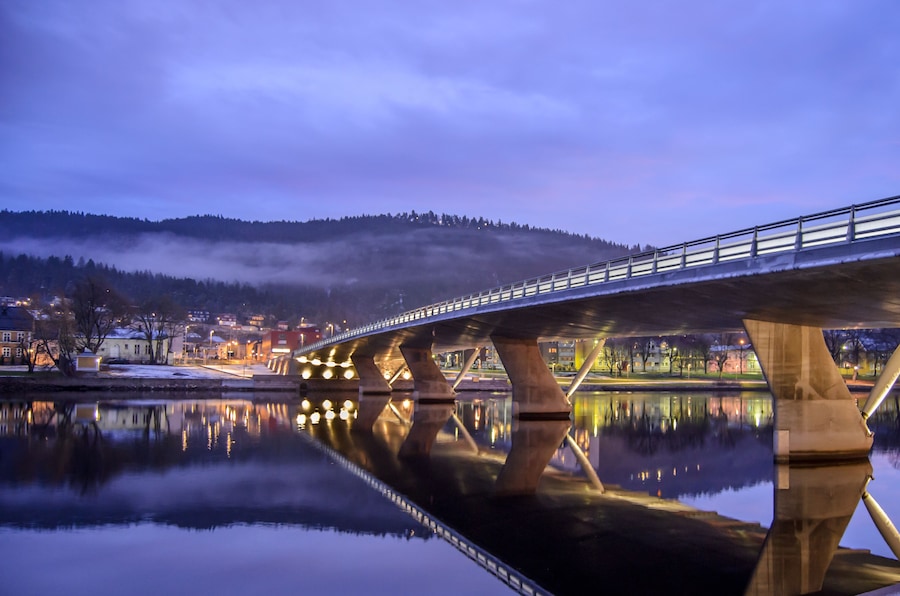 A bridge over a calm river reflecting the lights of a town and a mist-covered hill in the background.