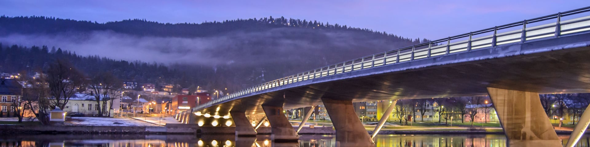 Uma ponte sobre um rio calmo que reflete as luzes de uma cidade e uma colina envolta em névoa ao fundo.