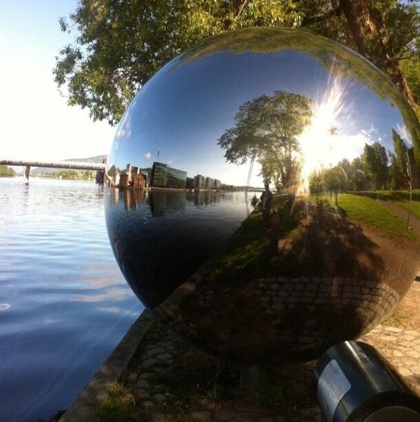 Uma esfera refletora grande em um parque, com um rio e uma ponte ao fundo.