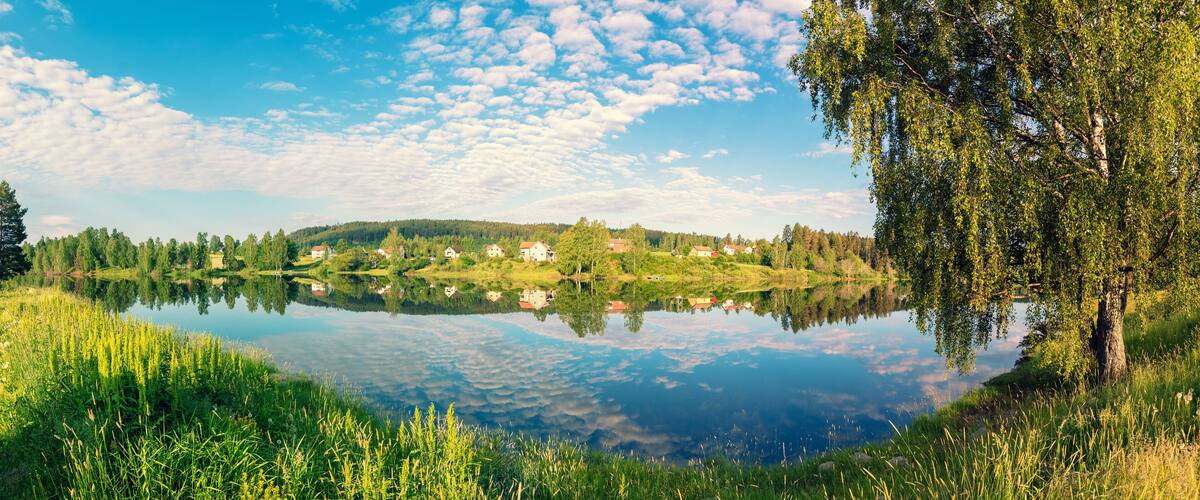 A calm lake with a reflection of houses and trees, surrounded by lush greenery and a clear sky.