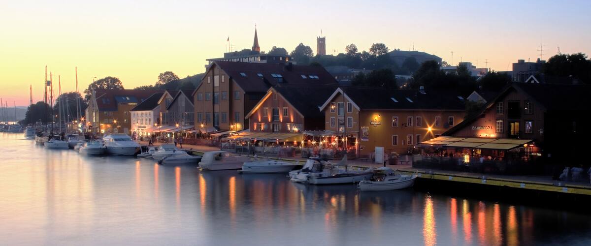 A harbor at dusk with boats docked, waterfront buildings, and a church spire in the background.