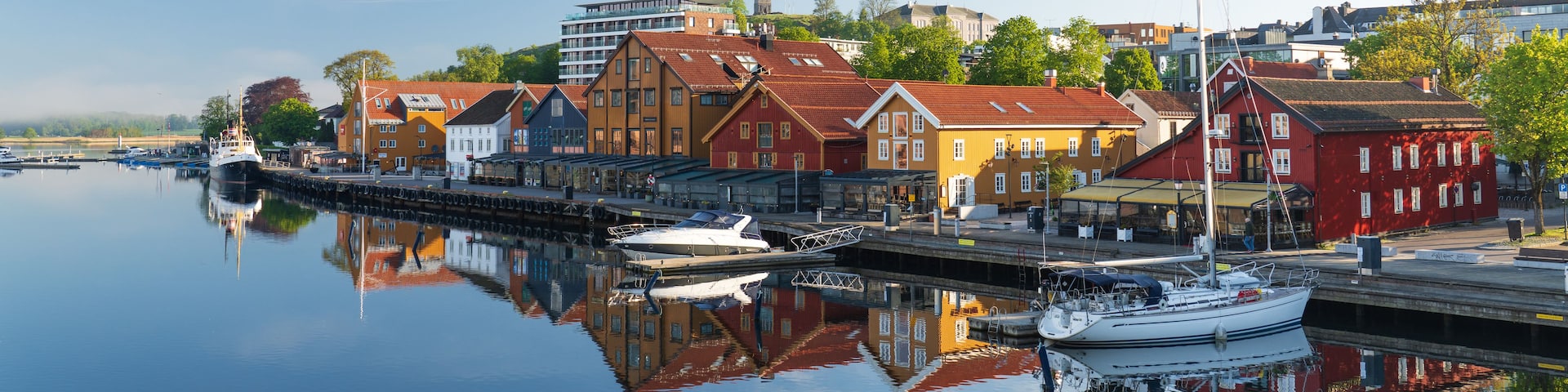 A calm harbor with a variety of buildings, including a church with a tall spire, and a modern building with a sloped roof.