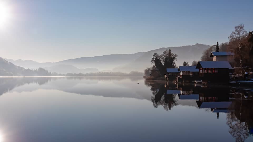 A calm lake with a clear reflection of buildings and trees, surrounded by misty mountains.