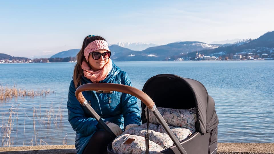 A person with a stroller by a calm lake with mountains in the background.