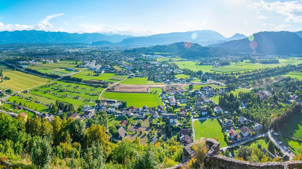 A panoramic view of a town nestled in a valley with surrounding mountains, green fields, and a river.