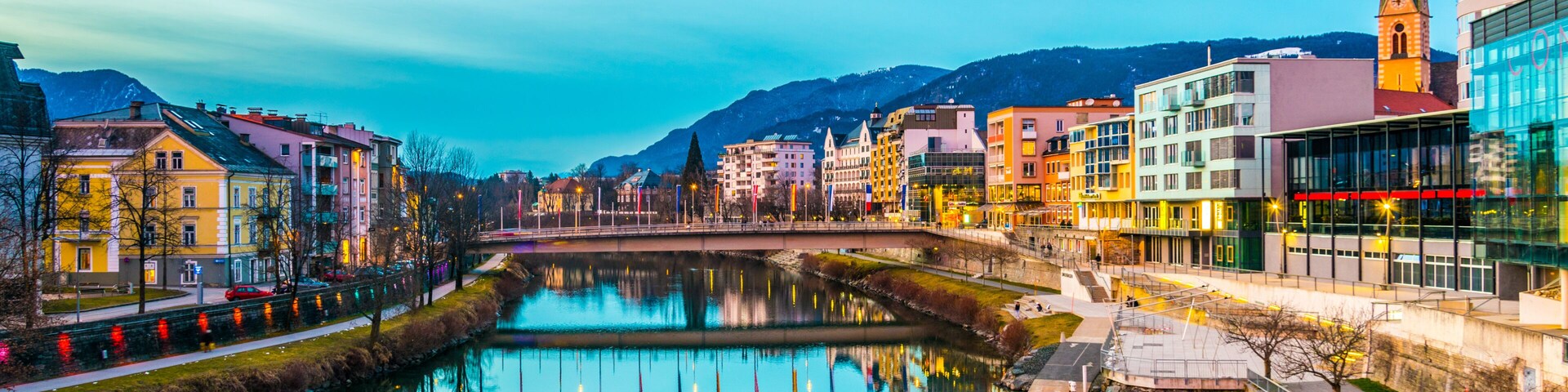 Um rio calmo refletindo edifícios, uma torre de igreja e uma montanha ao fundo.