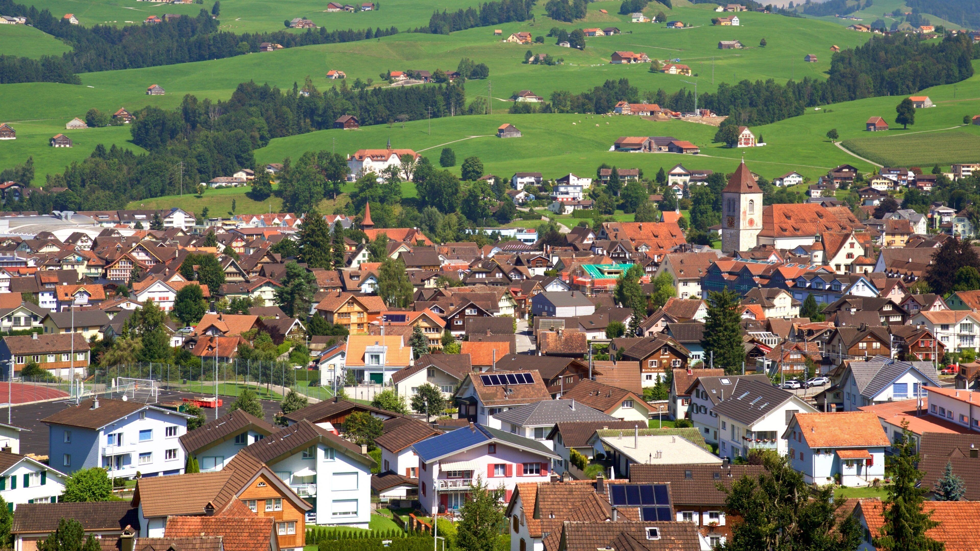 A densely populated village with traditional houses, a prominent church, and rolling green hills in the background.