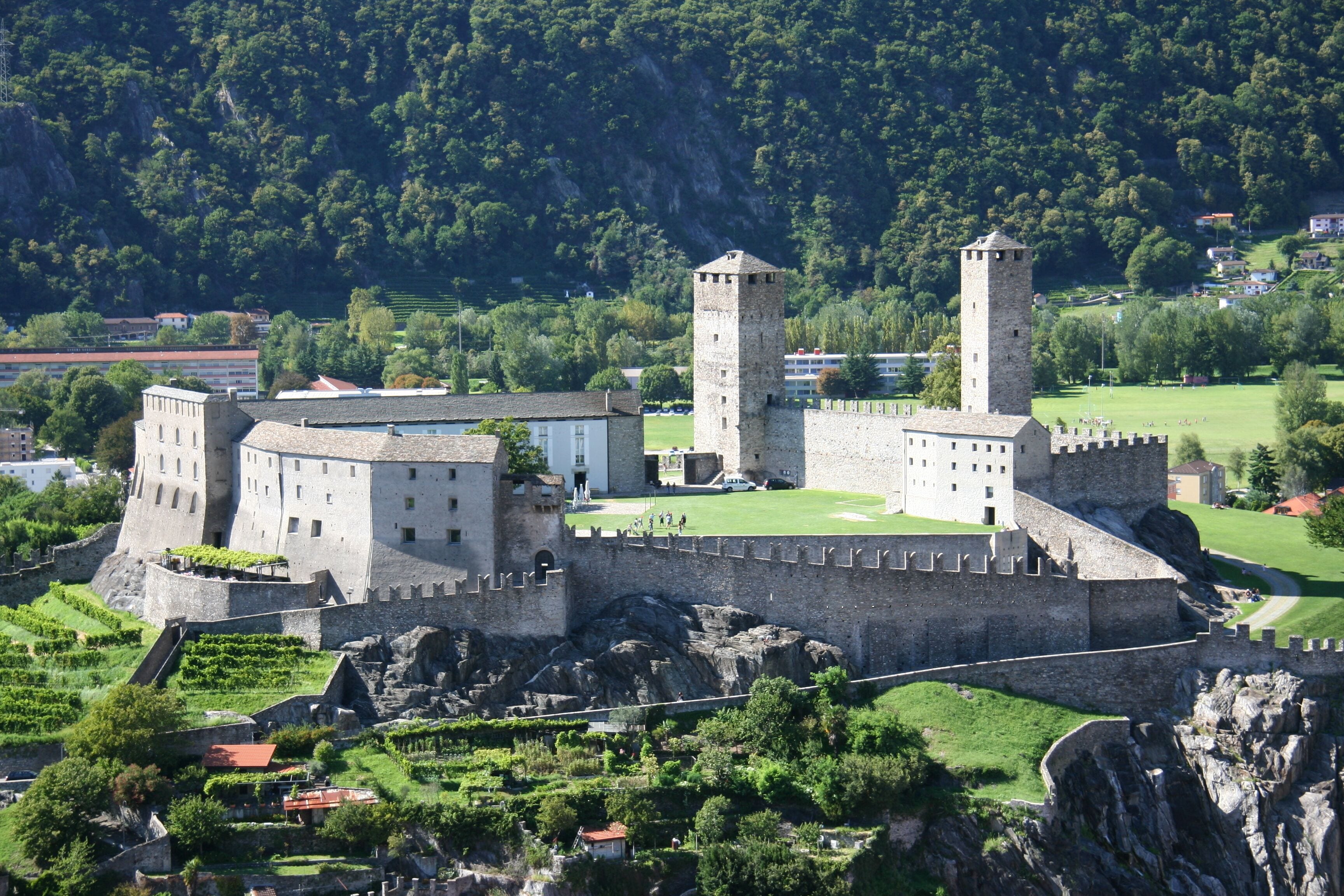 A historic castle with two towers, surrounded by lush greenery and a mountainous backdrop.