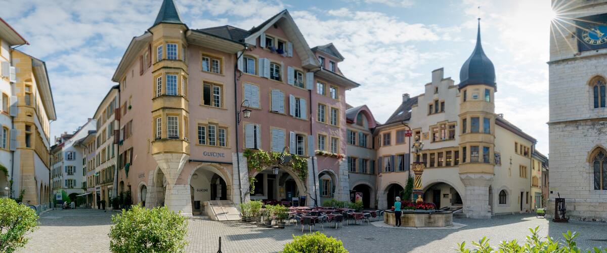 A European town square with historic buildings, a clock tower, and cobblestone streets.