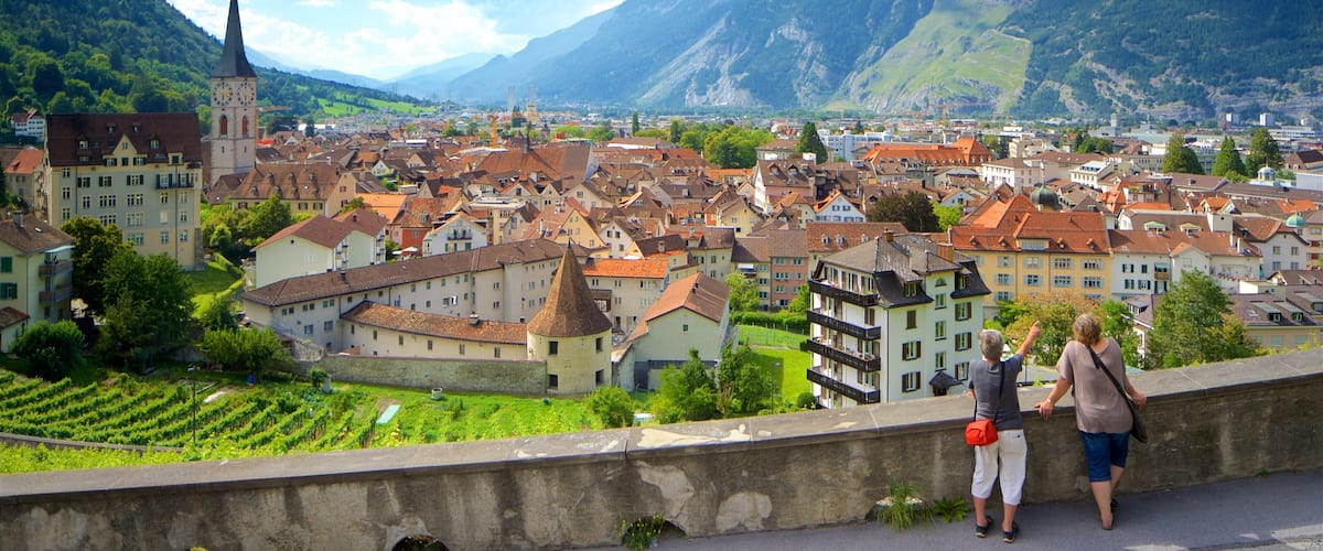A town with a prominent church steeple, surrounded by mountains and vineyards.