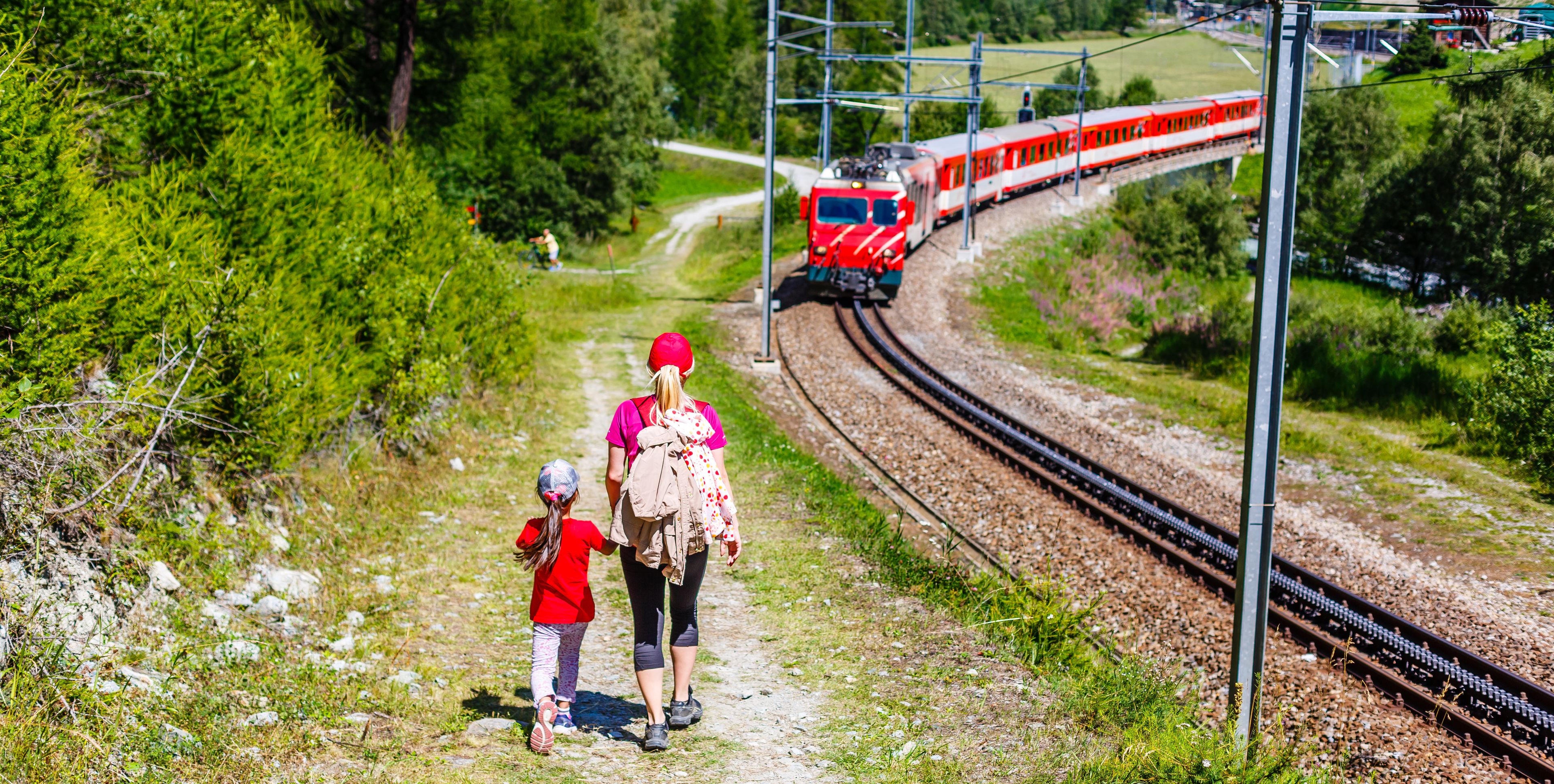 Uma mulher e uma criança caminham por um caminho ao lado de trilhos de trem, com um trem vermelho ao fundo.