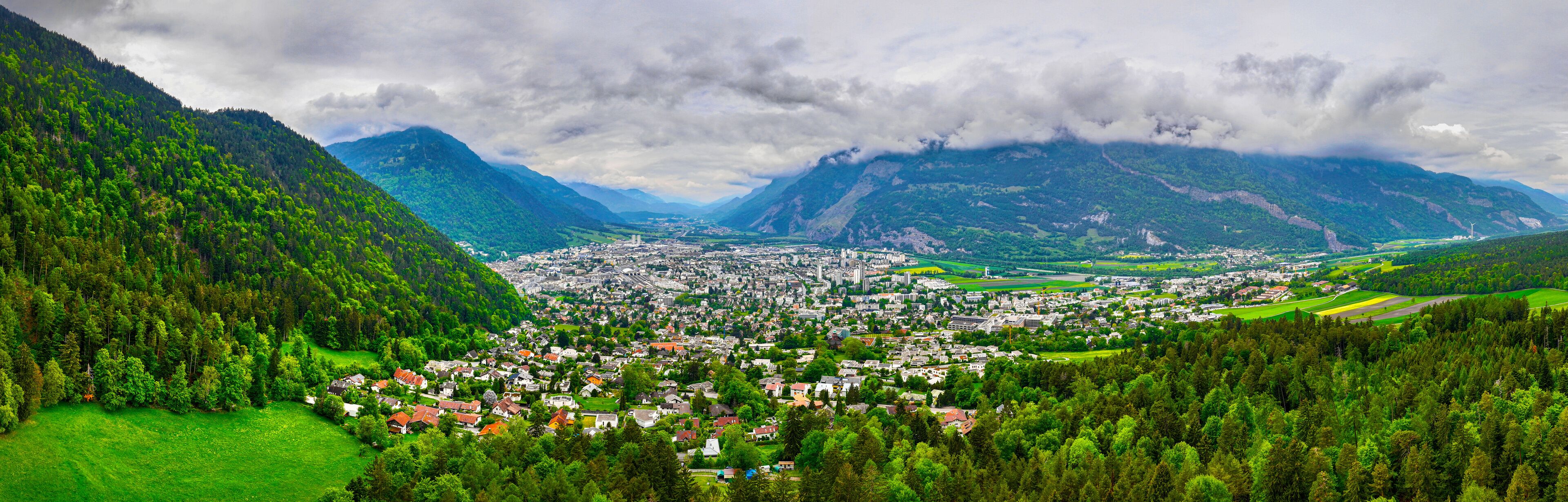 A town nestled in a valley surrounded by mountains, with lush greenery and a cloudy sky.