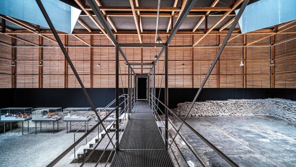 A modern architectural structure with a metal framework, wooden panels, and a central staircase leading to a dark doorway.
