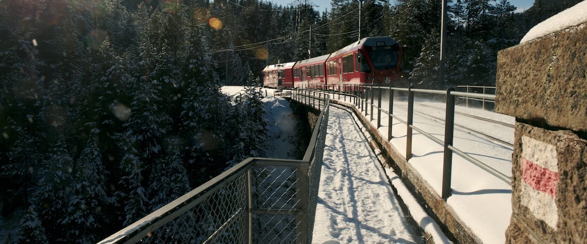 A red train on a snow-covered track, surrounded by snow-laden trees and a clear sky.