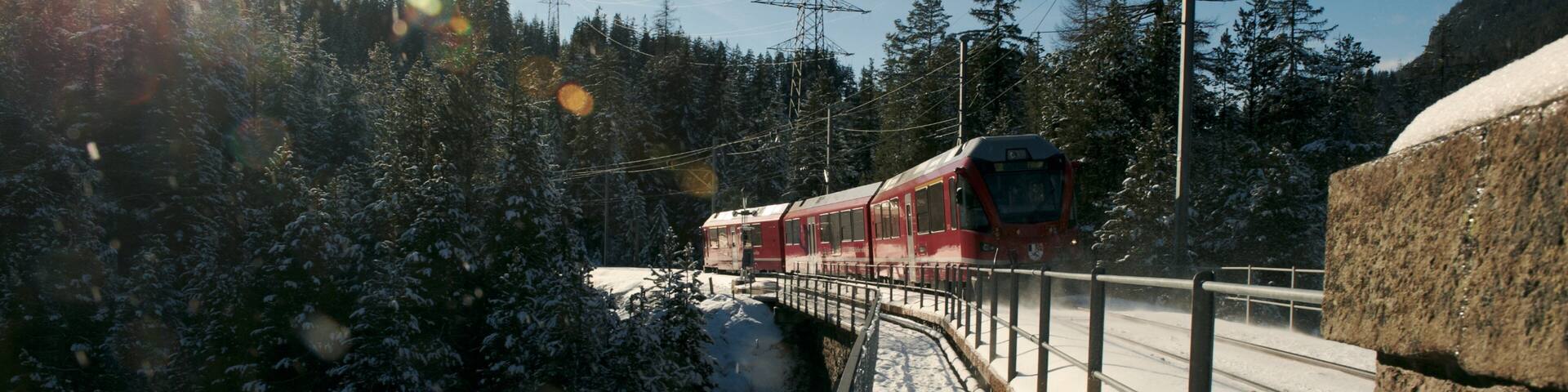 A red train on a snow-covered track, surrounded by snow-laden trees and a clear sky.