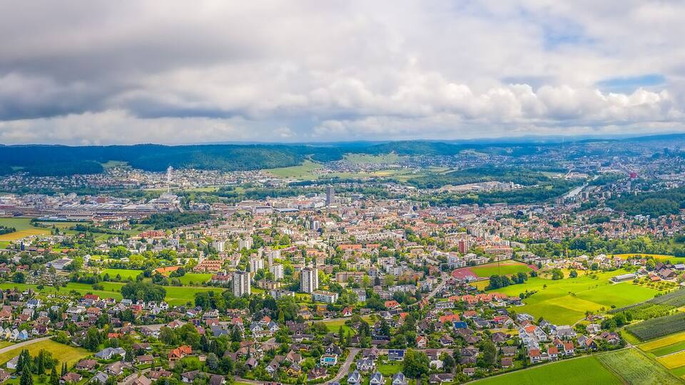 A panoramic view of a town nestled in a valley with surrounding hills, a river, and a distant mountain range.