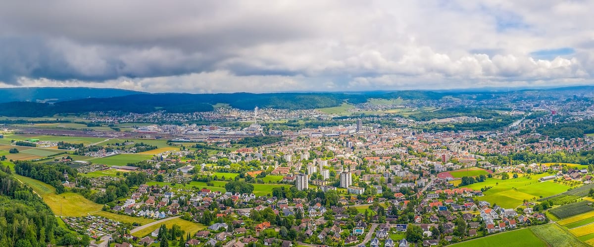 A panoramic view of a town nestled in a valley with surrounding hills, a river, and a distant mountain range.