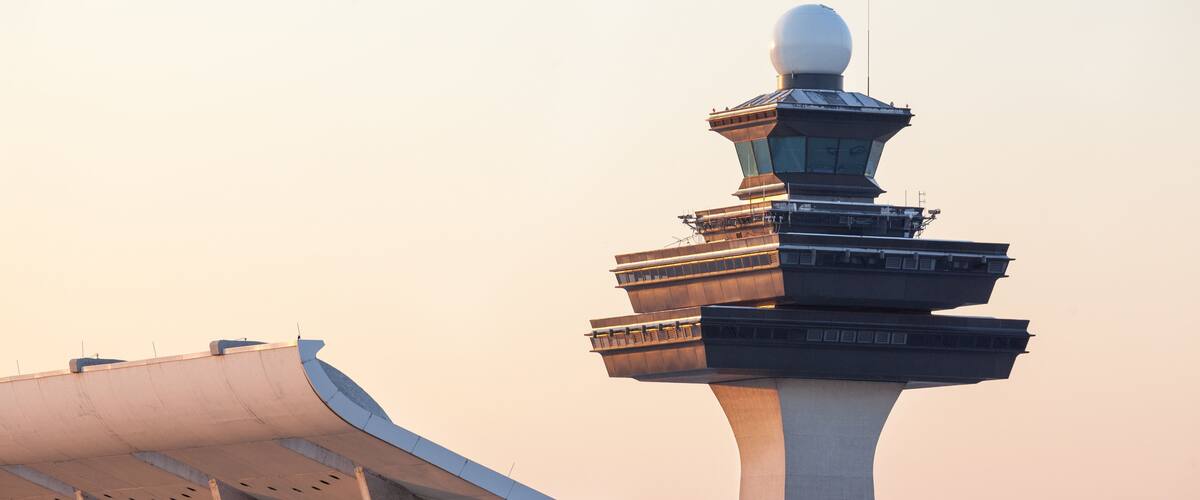 A modern airport control tower with a spherical top and a curved, white-roofed building in the foreground.