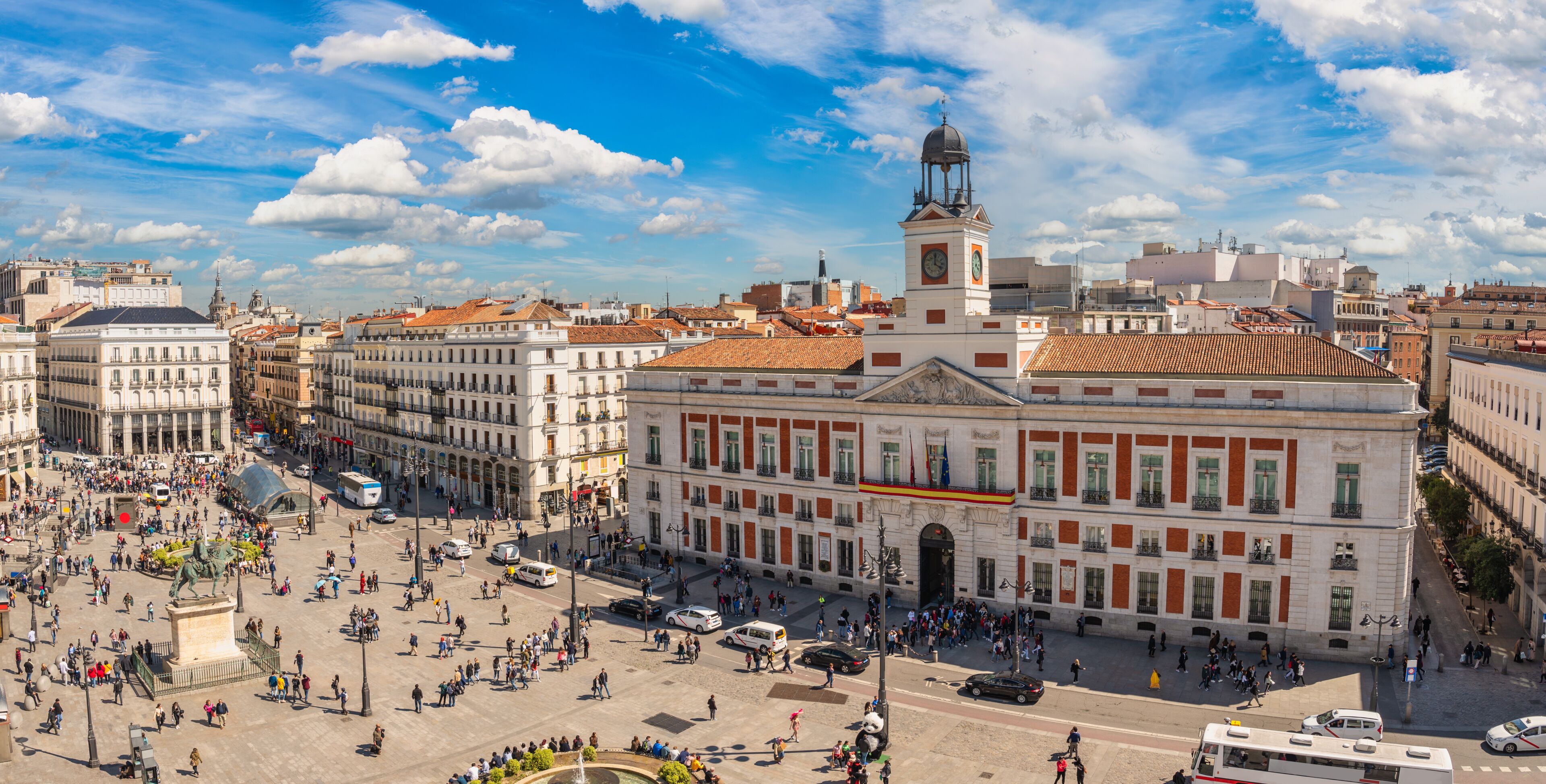 Una plaza animada con edificios históricos, una destacada torre del reloj y una estatua en el centro.