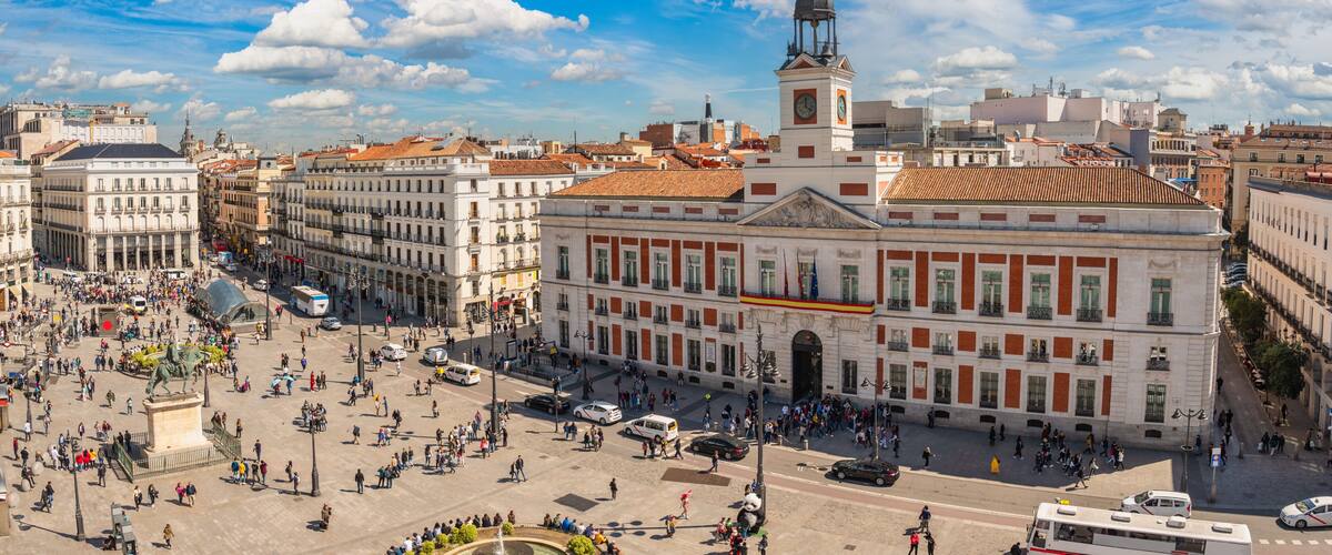 A bustling square with historic buildings, a prominent clock tower, and a statue in the center.