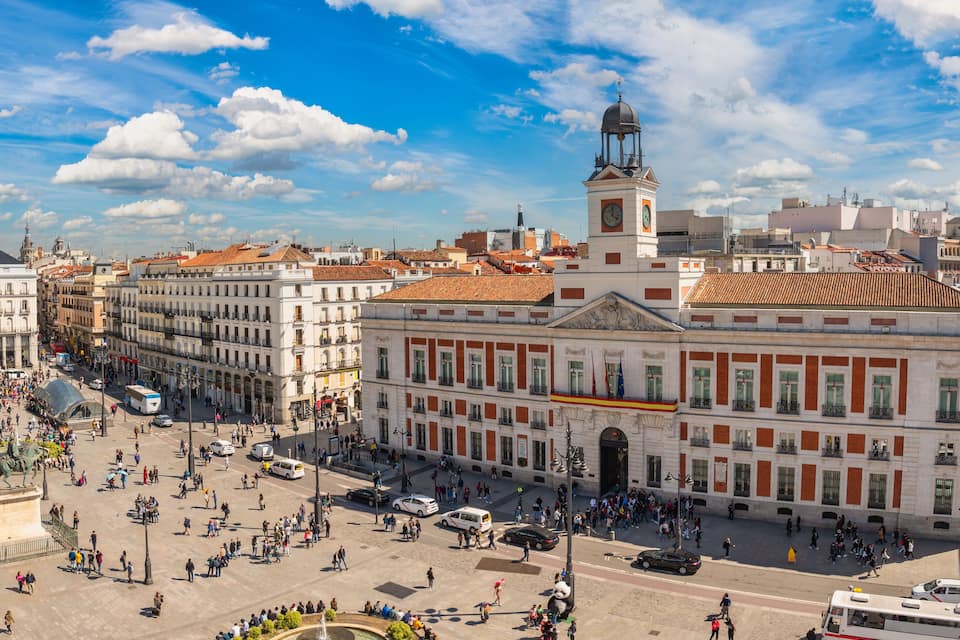 Una plaza animada con edificios históricos, una destacada torre del reloj y una estatua en el centro.