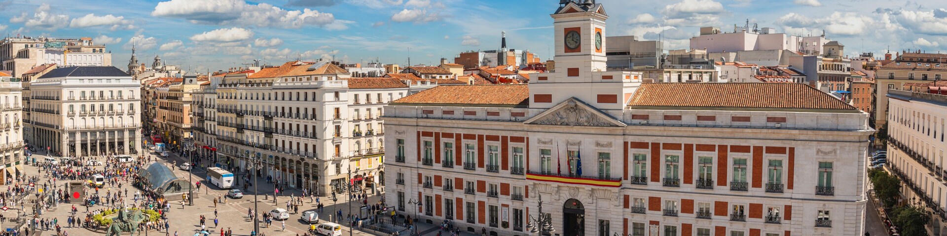 Uma praça movimentada com edifícios históricos, uma imponente torre do relógio e uma estátua no centro.