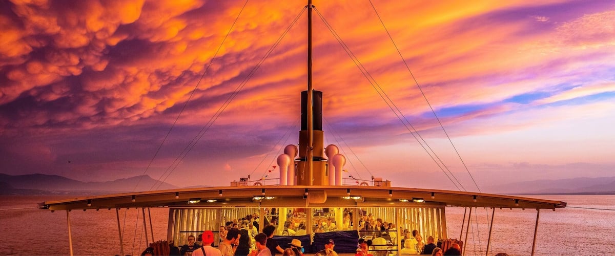 A ship with a large mast and a crowd of people on deck during sunset.
