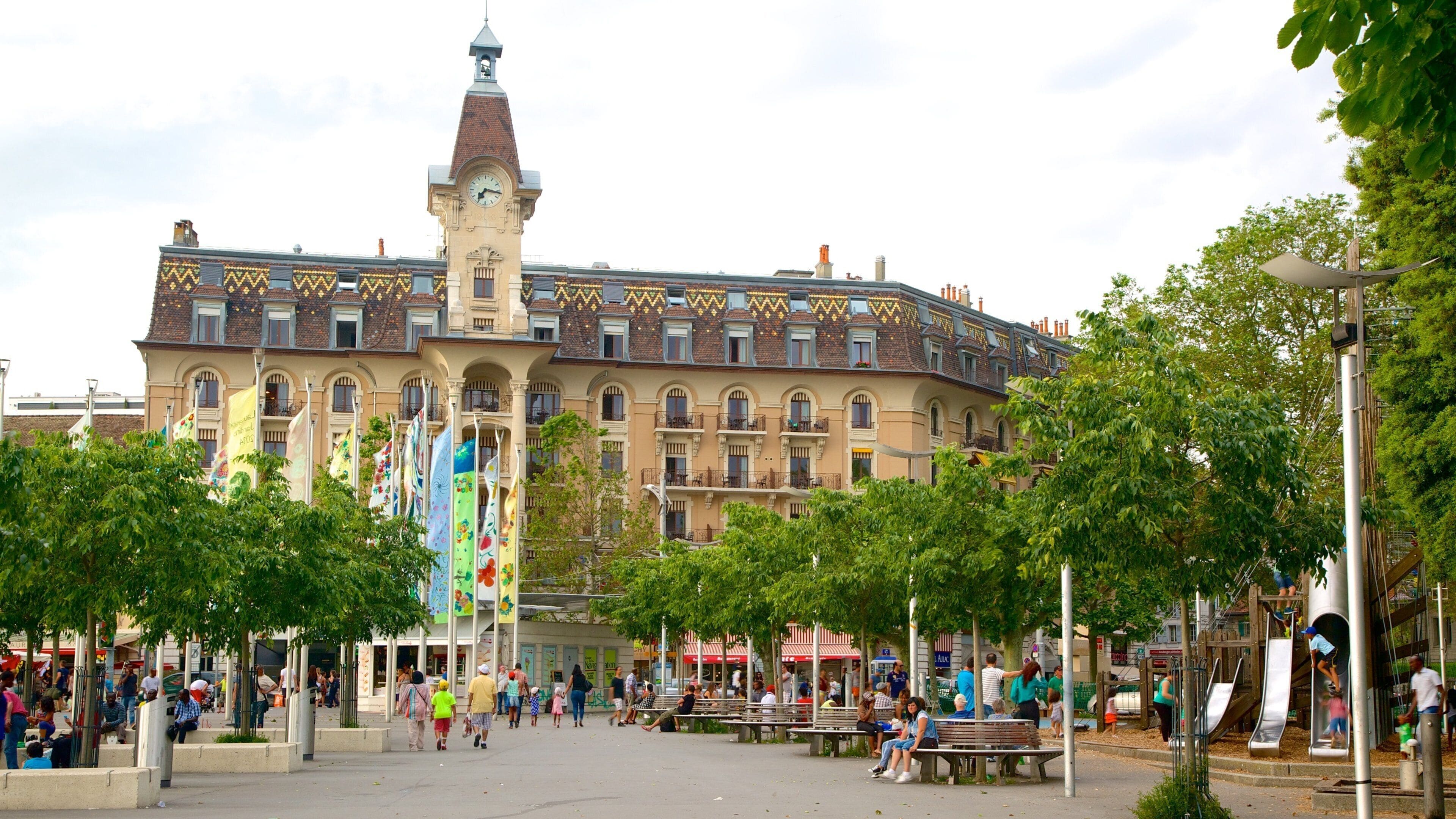 Un edificio histórico con una torre de reloj, rodeado de árboles y una plaza con bancos y gente.
