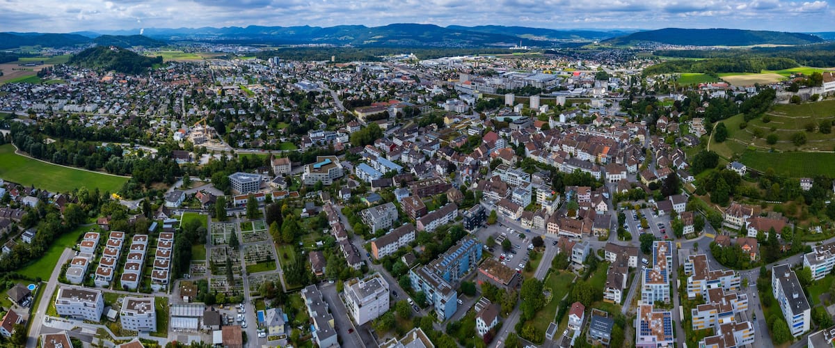 An aerial view of a densely populated city with a mix of residential and commercial buildings, surrounded by green fields and distant mountains.