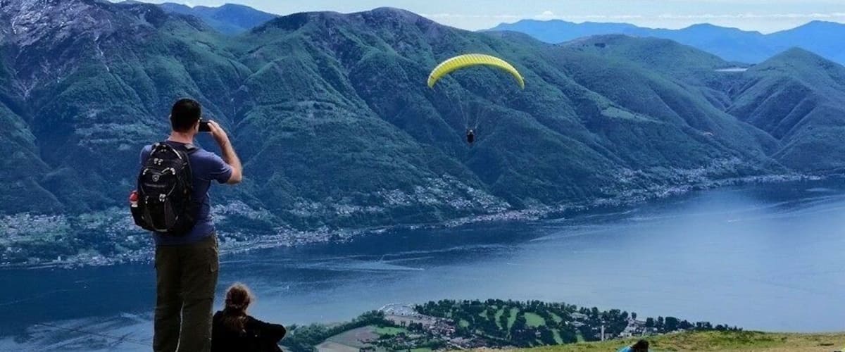 A person taking a photograph of a paraglider over a mountainous landscape with a lake below.