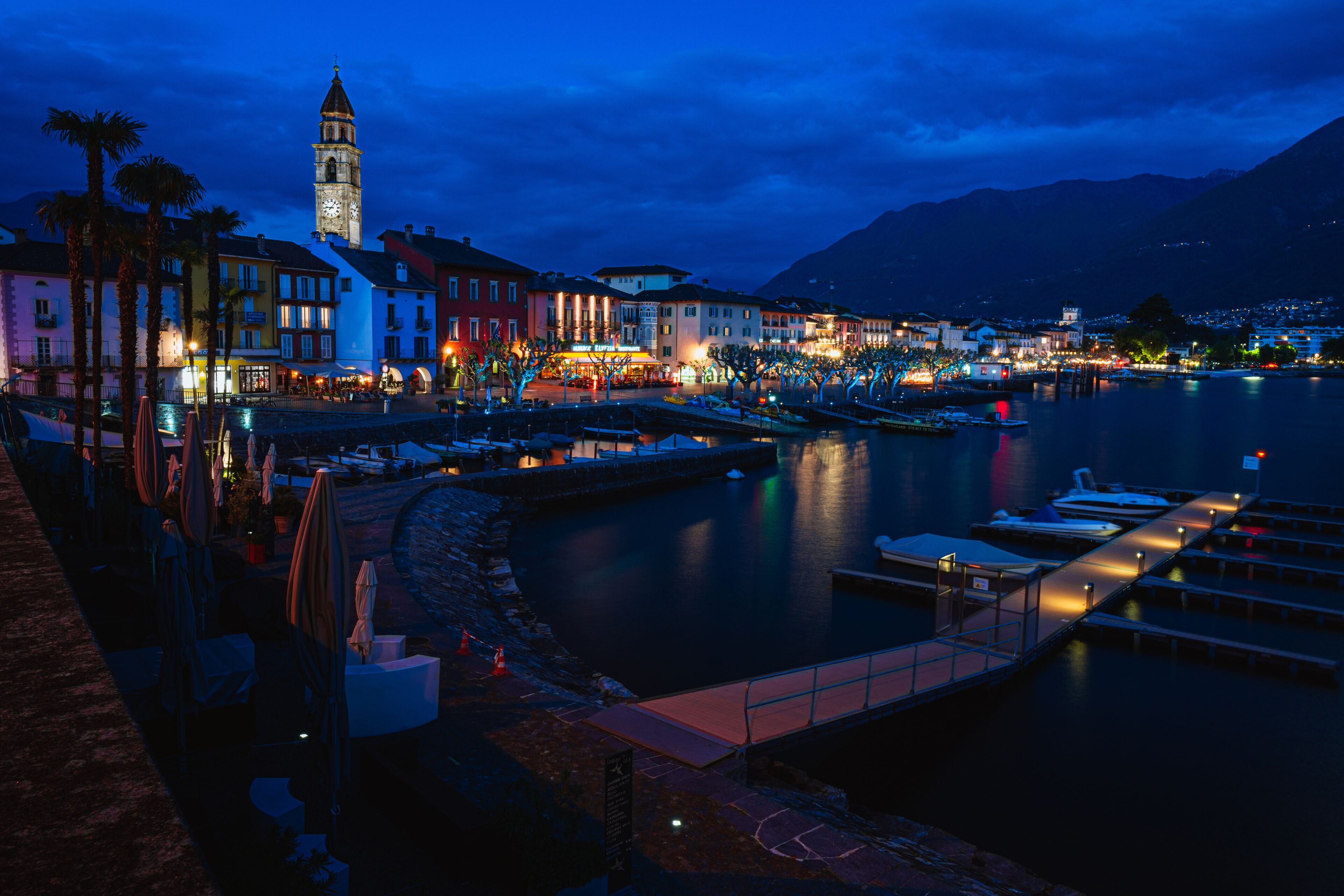Un pueblo frente al agua al anochecer, con edificios iluminados, una torre del reloj y un lago tranquilo.