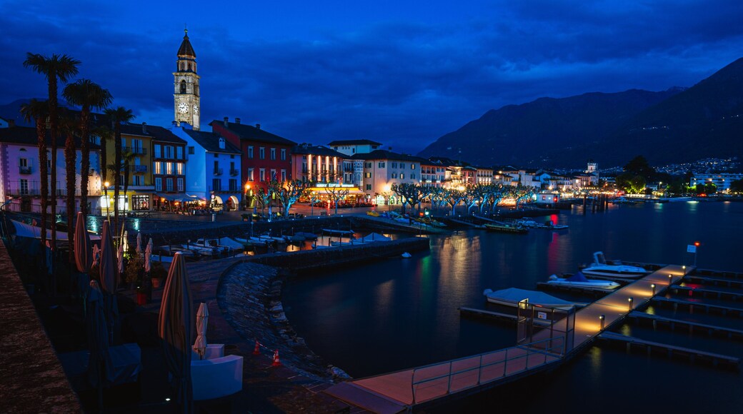 Un pueblo frente al agua al anochecer, con edificios iluminados, una torre del reloj y un lago tranquilo.