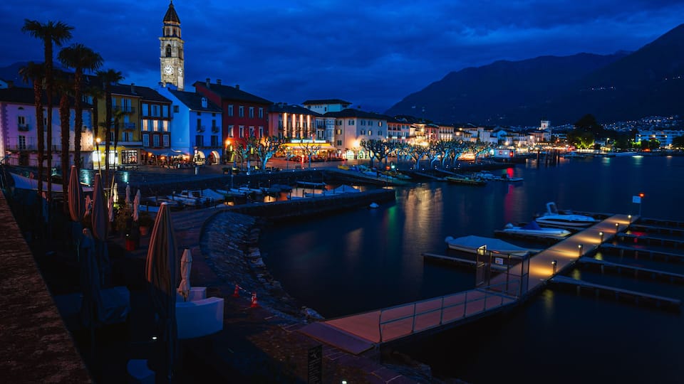 A waterfront town at dusk with illuminated buildings, a clock tower, and a calm lake.