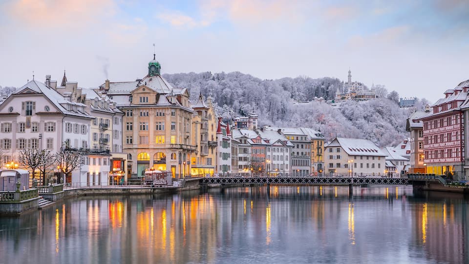 A riverside cityscape with historic buildings and a snow-covered hill in the background.