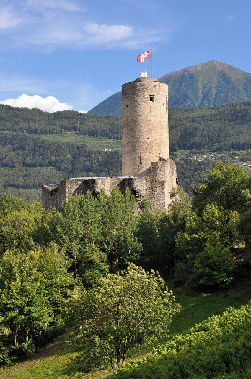 A historic stone tower with a flag, surrounded by lush greenery and mountains in the background.
