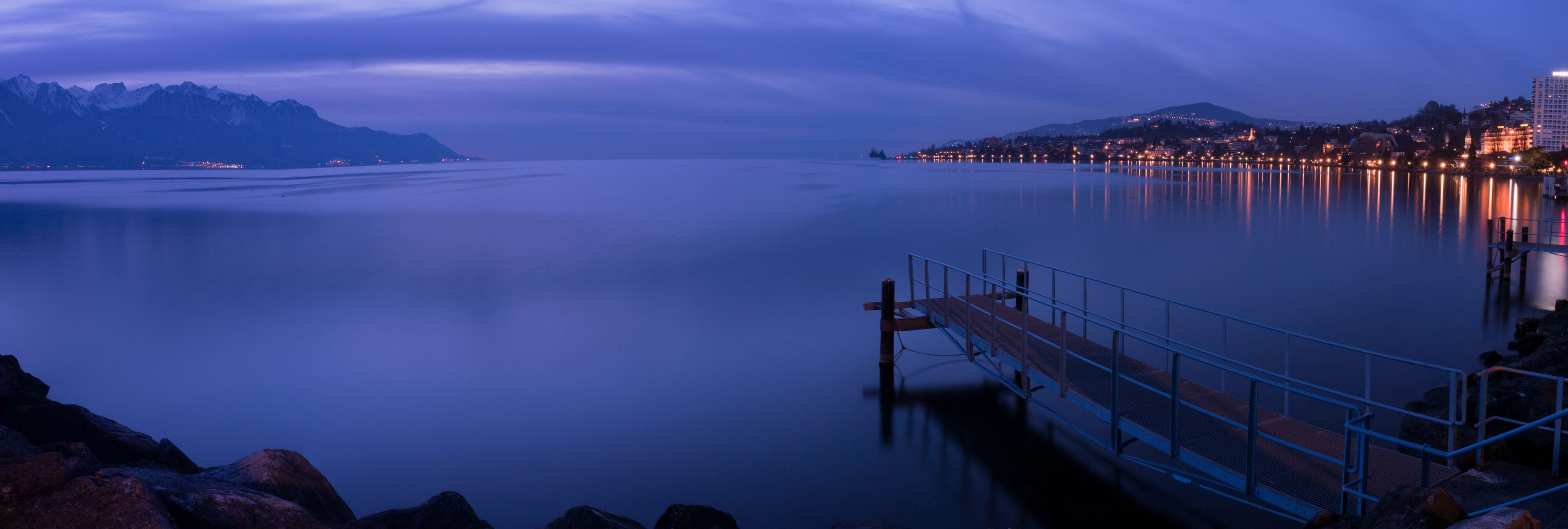 Un lago tranquilo al anochecer con un muelle, montañas al fondo y un paisaje urbano en la orilla opuesta.