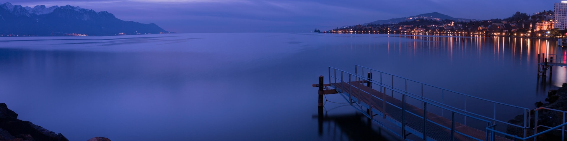 Un lago tranquilo al anochecer con un muelle, montañas al fondo y un paisaje urbano en la orilla opuesta.