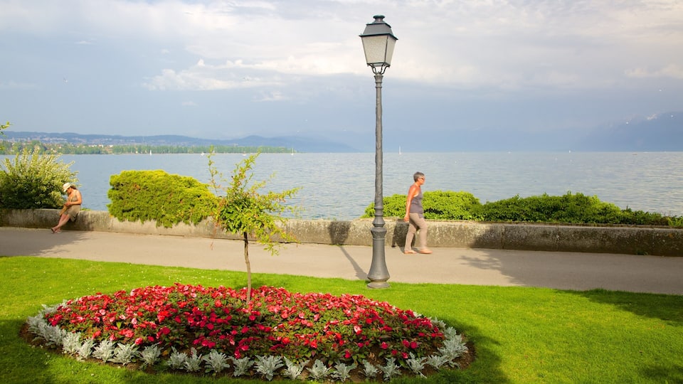 A lakeside walkway with a lamppost, red flowers, and a person walking.