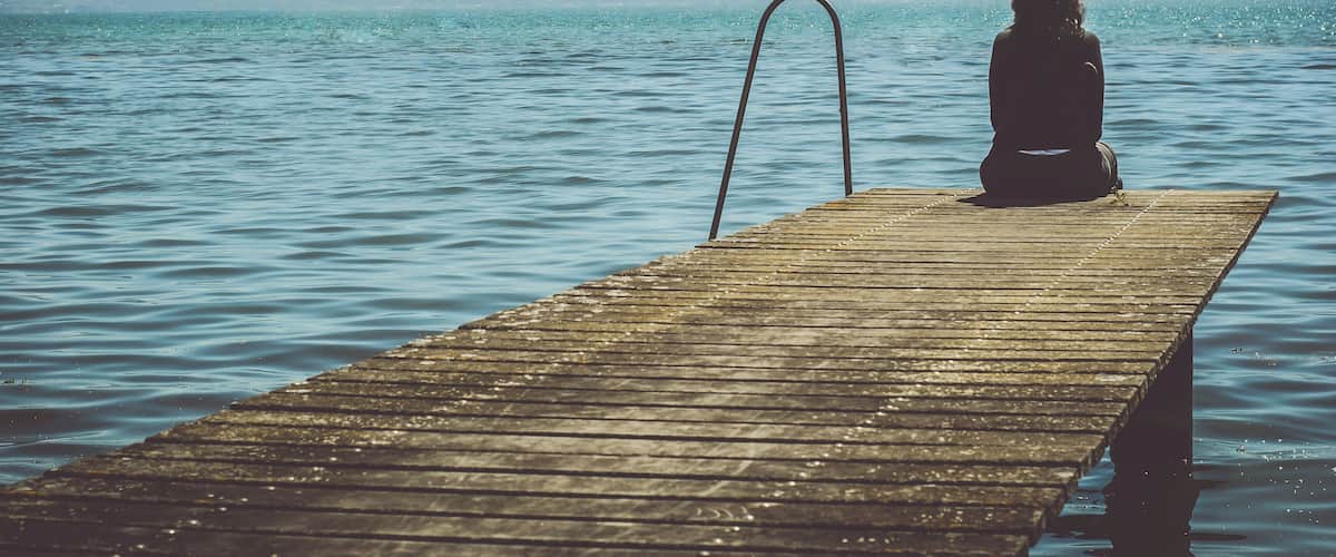 A person sitting on a wooden pier extending into a calm lake with mountains in the background.