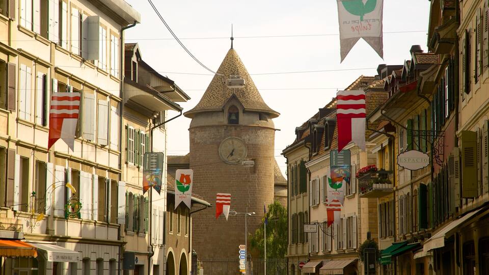 A narrow street with a prominent tower, buildings with green shutters, and flags.