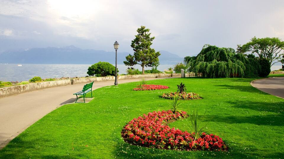 A park with a bench, flower beds, trees, a lamp post, and a view of the sea and mountains.