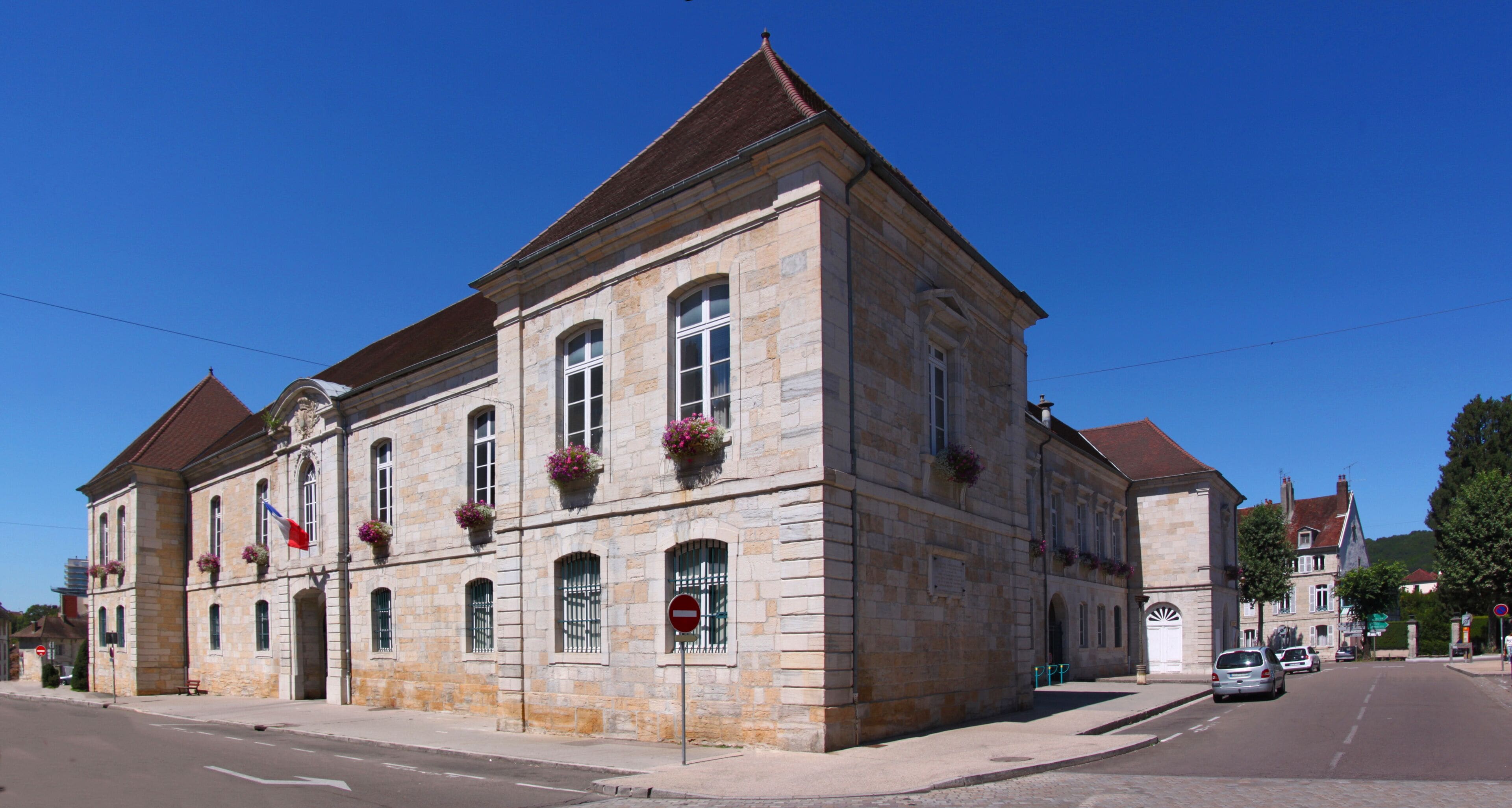 A historic stone building with arched windows and a red roof, situated on a sunny street.