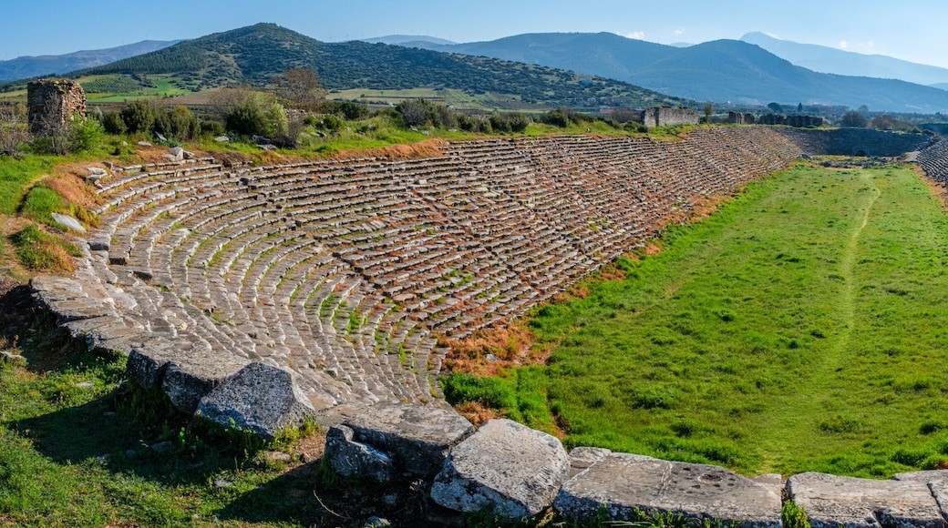 An ancient amphitheater with stone seating and a central grassy area, surrounded by hills and mountains.
