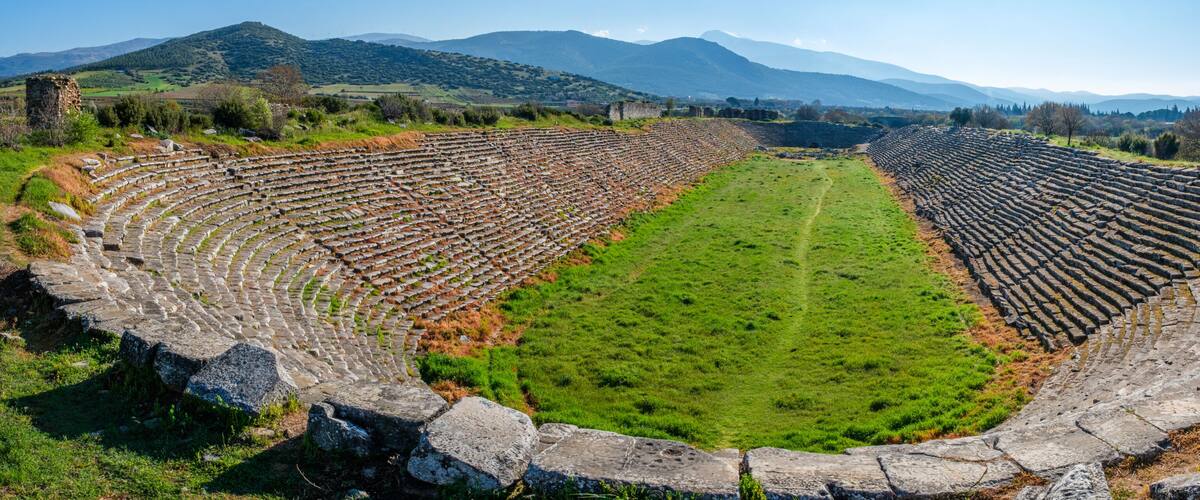 An ancient amphitheater with stone seating and a central grassy area, surrounded by hills and mountains.