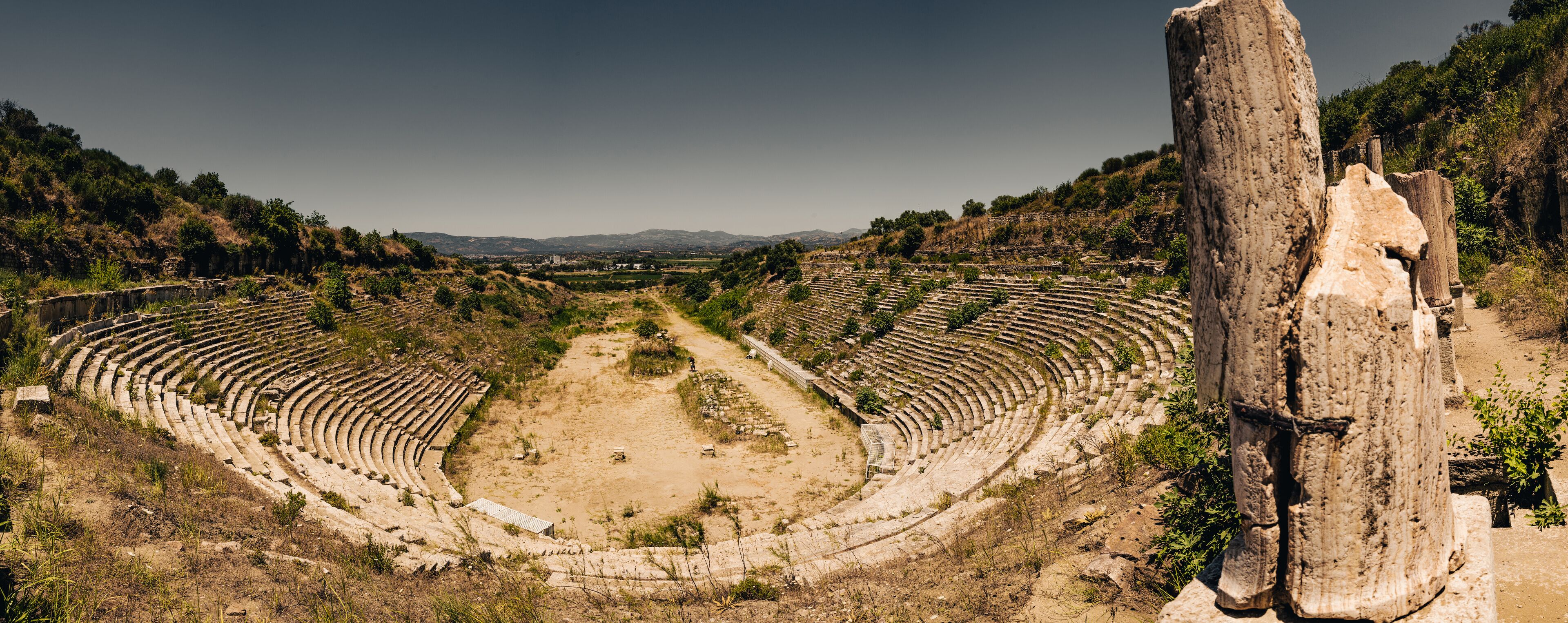 An ancient amphitheater with tiered seating and a central stage, surrounded by hills and vegetation.